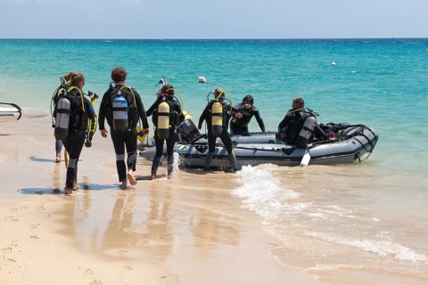 Recreational divers walk across beach to inflatable boat in diving suits with scuba tanks on their