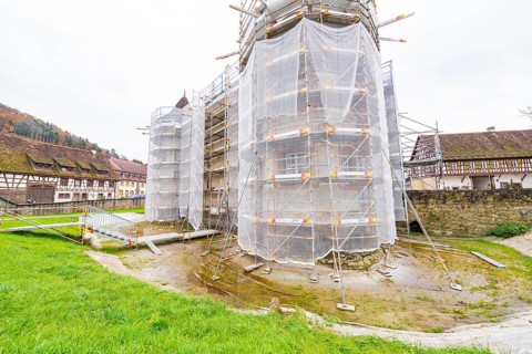A rural-style renovated building with scaffolding and tarpaulins, surrounded by green spaces,