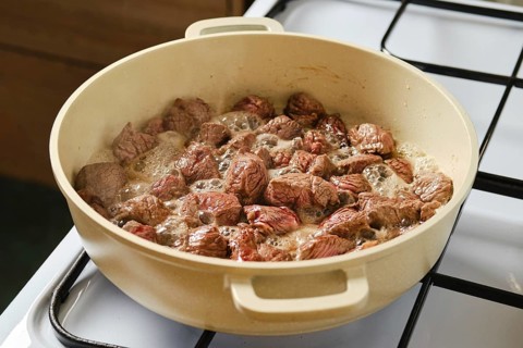 Pieces of beef are sizzling and cooking in a large pot on a gas stove. The meat is browning while