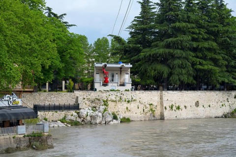 A riverbank cable car station surrounded by lush trees and adjacent buildings, cable car cabin,