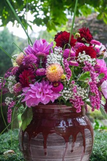 A vivid bouquet of flowers in shades of pink and red stands in a rustic vase against a green
