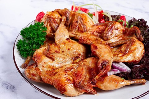 Baked quail, served on a plate with crispy lettuce leaves, fresh tomatoes and herbs, on a white