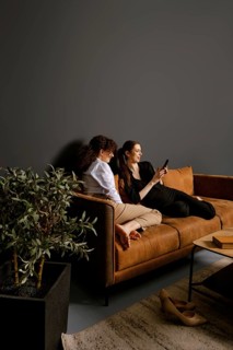 Two friends sit comfortably on a brown couch in a modern living room, engaged with a mobile device.
