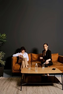 In a stylish indoor setting, two individuals sit on a brown couch. One person reads a book while