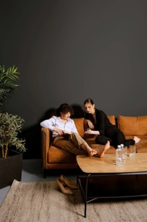 Two friends sit comfortably on a warm brown couch in a stylish room, engaging in conversation. Lush