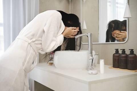 A woman in a white bathrobe leans over a stylish sink in a contemporary bathroom, splashing water