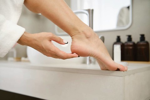 A person gently massages their foot while resting on a sink in a stylish bathroom. Soft morning