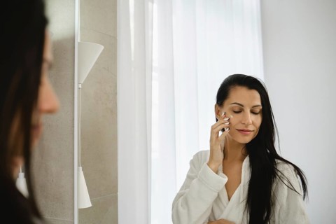 In a modern bathroom filled with natural light, a woman in a white robe applies skincare to her