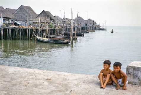 Two boys sitting on quayside next to attap houses of fishing village on stilts, Singapore,