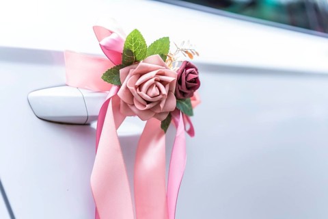 Elegant pink ribbons and artificial flowers adorn the handle of a white car, signifying a wedding