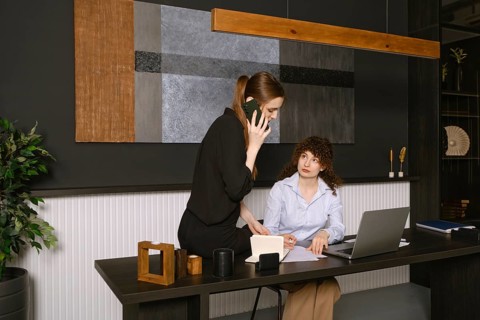 Two women are engaged in a work discussion in a stylish office setting. One is seated at a desk
