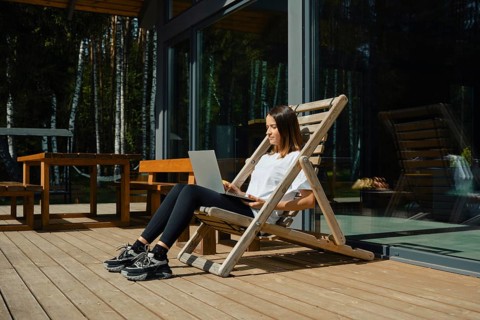 A woman sits comfortably in a wooden chair on a deck, focused on her laptop. Bright sunlight