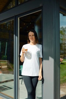 A young woman stands leisurely at the entrance of a stylish house, holding a drink in her hand.