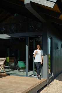 A woman stands on the wooden deck of a stylish cabin, holding a drink while enjoying the serene