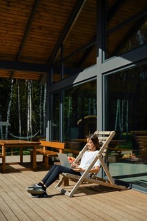 A woman sits on a wooden chair outdoors and typing on a laptop. She enjoys her vacation in modern
