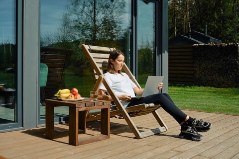 A woman sits comfortably in a deck chair on a wooden patio. She opened the laptop cover to check