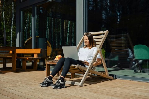 A woman is sitting in a wooden lounge chair, engaged with her laptop on a sunny deck. She enjoys a