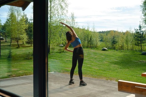The view from inside the house at a woman is stretching her body on a wooden deck surrounded by