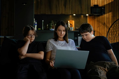 A woman and two children, are sitting closely on a sofa. They focus on a laptop screen in a warmly