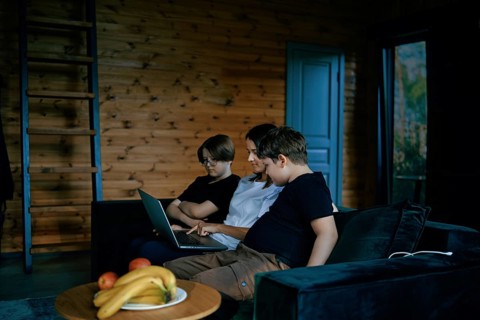 A family sits together on a comfortable couch in a wooden living room, focusing on a laptop. The
