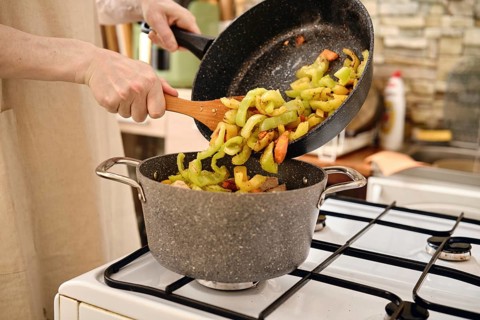 A person transfers sliced vegetables in a pot while adding more ingredients from a frying pan on a