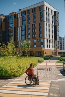 A man using a wheelchair navigates a crosswalk in a modern urban setting. The sun shines brightly,