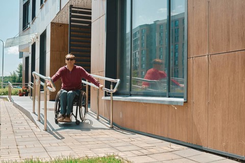 Individual in a wheelchair is utilizing a ramp to exit a modern building with large windows. The