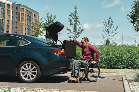 A person in a wheelchair is carefully placing a suitcase into the trunk of a parked car. The