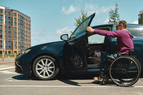 A man in a wheelchair approaches a car parked in a bright urban setting. He is wearing a purple
