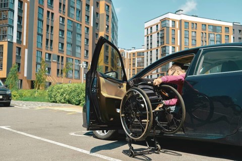 A person is maneuvering a wheelchair into a car parked in a contemporary urban area. A man unfolds