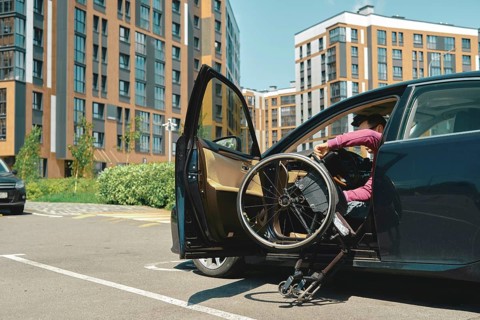A person is carefully getting out a vehicle using a wheelchair in a bustling parking lot. The