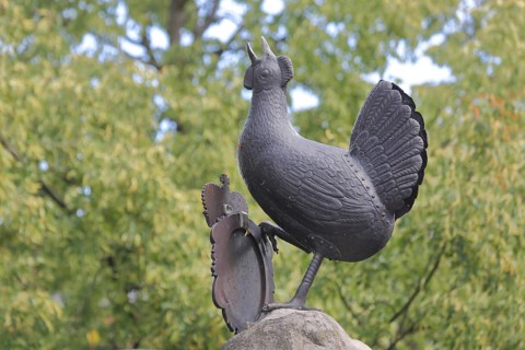 Grey bronze sculpture at the Hennebrunnen fountain, bird figure, screaming, tail feathers, market