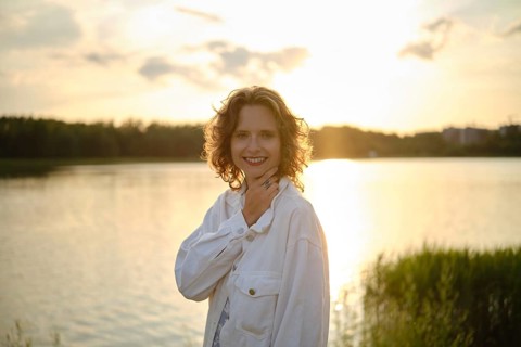 A woman stands by the lake, smiling as the sun sets behind her, casting a warm golden glow on the