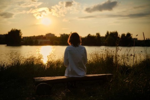 At the edge of a serene lake, a woman sits on a wooden bench surrounded by tall grass, soaking in