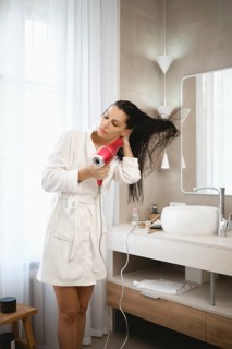 A woman stands in a modern bathroom, using a hair dryer to style her long, wet hair. She is dressed
