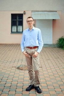 A man stands confidently outside a modern building, wearing a light blue shirt and beige pants. His
