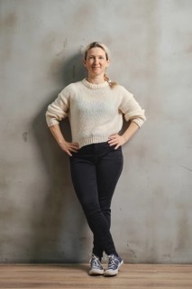 A woman stands casually with her hands on her hips, wearing a soft sweater and dark jeans. She