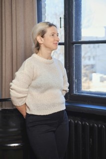 A woman with light hair is standing by a window, gazing thoughtfully outside. She is dressed in a