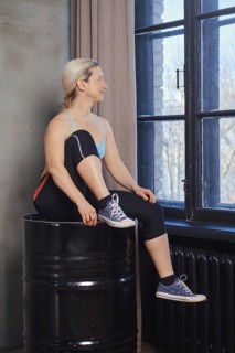 A woman sits on a barrel, pausing between exercises. She gazes out the large windows, showcasing