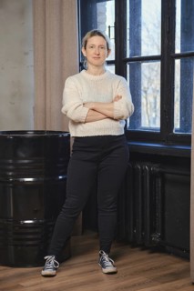 A woman stands with arms crossed, dressed in a white sweater and dark pants, exuding confidence in