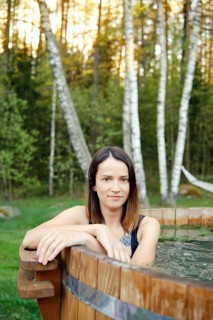 A woman enjoys a tranquil moment in a wooden hot tub, framed by tall trees and lush greenery during