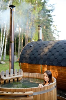 A woman enjoys a peaceful moment in an outdoor wooden hot tub surrounded by tall trees. Steam rises