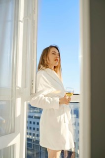 A woman stands on a balcony, dressed in a soft white robe, sipping a glass of beverage. She gazes