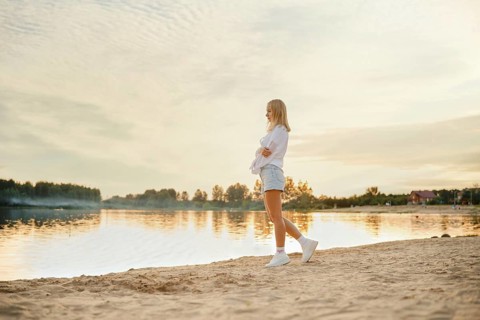 A young woman explores a sandy beach as the sun sets over a calm lake. The scenery features