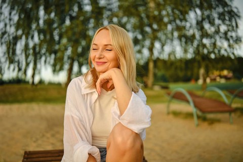 A pleased woman sits comfortably on a bench by the lake, smiling softly as the sun sets