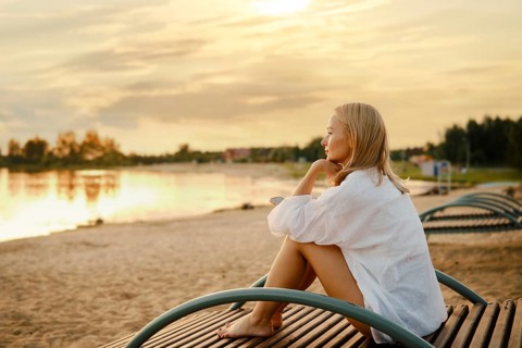 A woman sits on a wooden bench by the shore, gazing at a stunning sunset over the water. The soft