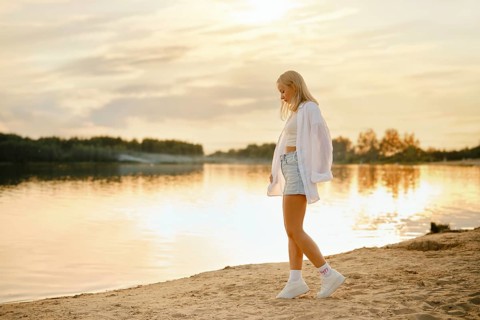 A young woman strolls on the sandy beach at sunset, with the golden light reflecting on the calm