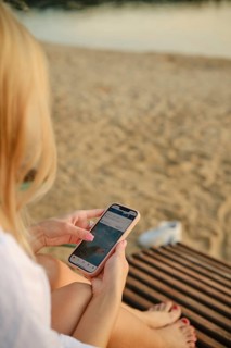 A woman sits on a wooden lounge, absorbed in her smartphone. The sun sets in the background,