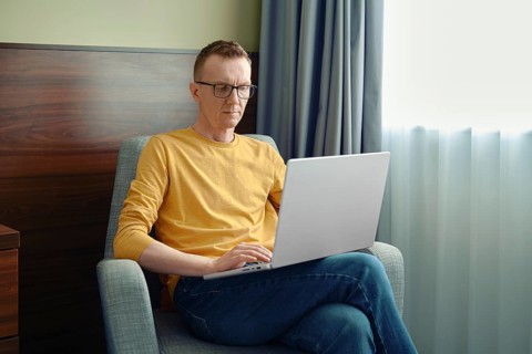 A man sits in a comfortable chair, focused on his laptop in a bright room. Soft natural light