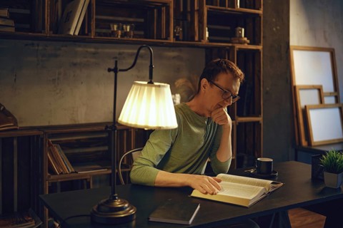 A man sits at a desk in a dimly lit room, engrossed in a book. Warm light from a lamp highlights